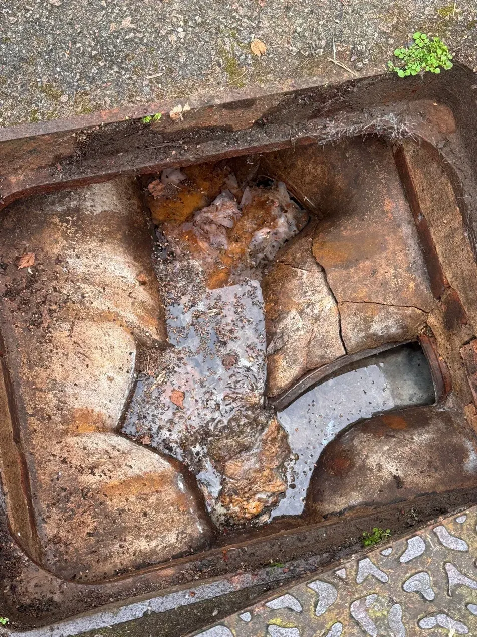 Before clearing, a cracked roadside gully chamber filled with standing water, soggy wipes and compacted debris.